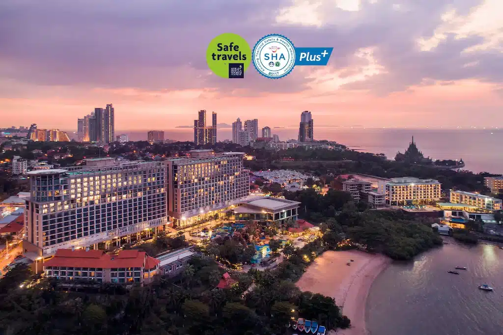 มุมมองทางอากาศของเมืองชายฝั่งยามพระอาทิตย์ตกดินพร้อมอาคารสูง ริมชายหาด และโลโก้รับรองสำหรับ Safe Travels และ SHA Plus+ ที่ตรงกลางด้านบน เน้นตัวเลือกพัทยาราคาถูก.