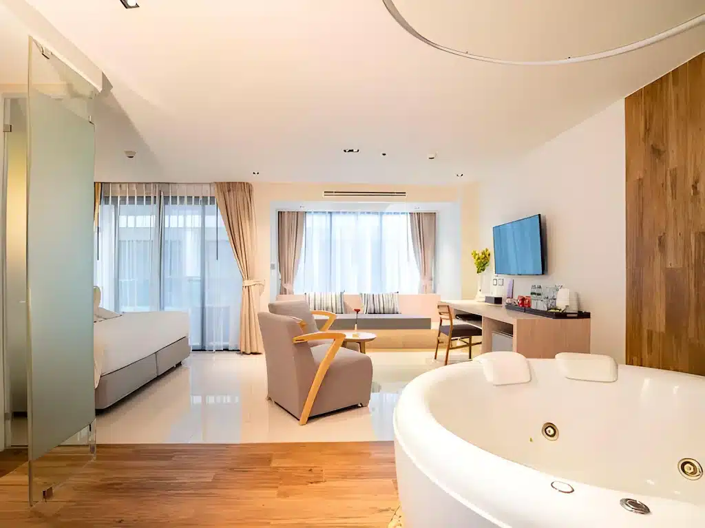 ห้องสวีทโรงแรมทันสมัยพร้อมเตียง พื้นที่นั่งเล่น หน้าต่างบานใหญ่พร้อมผ้าม่าน ทีวีจอแบน และอ่างจากุซซี่ทรงกลมอยู่เบื้องหน้า เหมาะสำหรับผู้ที่กำลังมองหาบริการพัทยาราคาถูกอย่างมีสไตล์.