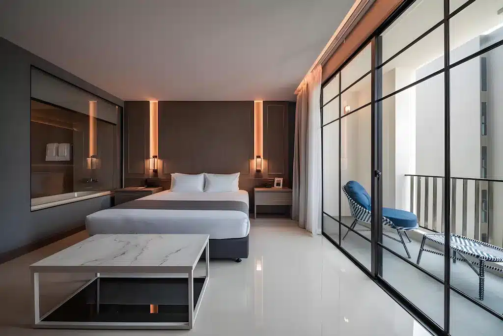 ห้องพักในโรงแรมทันสมัยพร้อมเตียงนอนที่จัดอย่างประณีต การตกแต่งแบบเรียบง่าย โต๊ะกาแฟหินอ่อน หน้าต่างบานใหญ่ และระเบียงพร้อมเก้าอี้นั่งเล่น 2 ตัว เป็นตัวเลือกที่เหมาะสำหรับที่พักในพัทยาที่แสวงหาความสะดวกสบายและมีสไตล์.