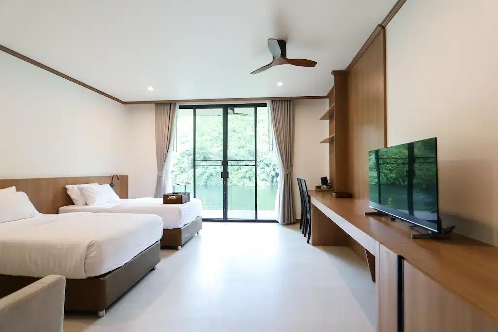 ห้องพักโรงแรมสมัยใหม่ในรีสอร์ทกาญจนบุรี พร้อมเตียงเดี่ยวสองเตียง, โทรทัศน์ติดผนัง, โต๊ะทำงานยาว, และประตูบานใหญ่เปิดออกสู่ ที่พักกาญจนบุรี ธรรมชาติสีเขียวภายนอก.