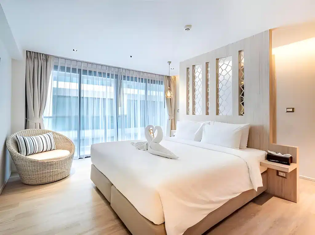 ห้องพักโรงแรมทันสมัยในที่พักพัทยาพร้อมเตียงขนาดใหญ่ ผ้าขนหนูรูปหงส์ เก้าอี้หวาย หน้าต่างสูงจากพื้นจรดเพดานพร้อมผ้าม่านโปร่ง และเฟอร์นิเจอร์ไม้สีอ่อน.