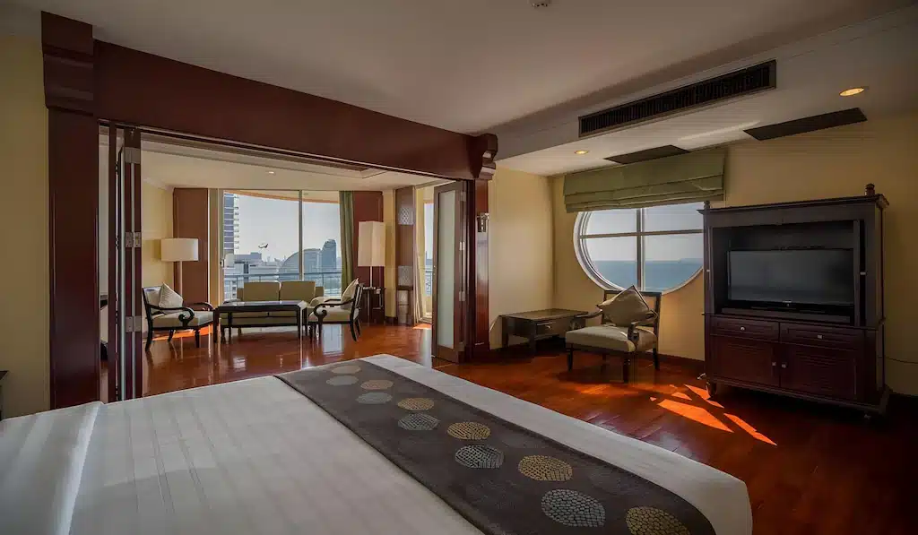 ห้องพักในโรงแรมที่มีเตียงขนาดใหญ่ พื้นที่นั่งเล่นริมหน้าต่างที่มองเห็นวิวทะเล พื้นไม้ ตู้วางทีวี และเฟอร์นิเจอร์ที่ทันสมัย เหมาะสำหรับผู้ที่กำลังมองหาพัทยาราคาถูกที่มีสไตล์และความสะดวกสบาย.