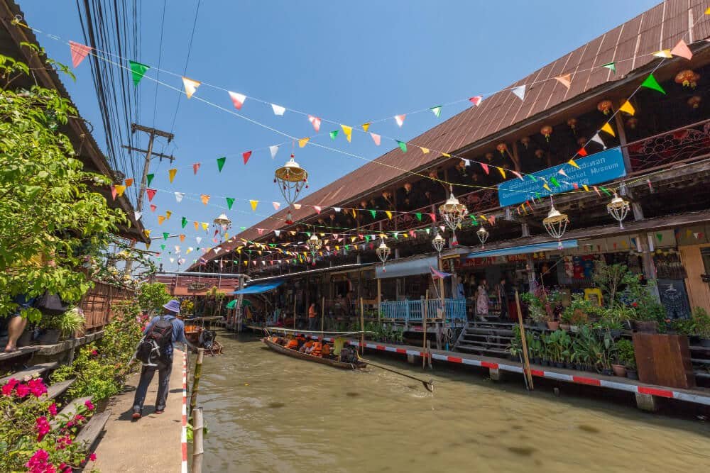 ตลาดริมคลองและธงสีสันสดใส เที่ยวใกล้กรุงเทพ ต้อนรับผู้มาเยือนสู่สถานที่คึกคักแห่งนี้—หนึ่งในสถานที่ท่องเที่ยวที่ดีที่สุดในสตูล—ซึ่งเรือแล่นผ่านร้านค้าที่เรียงรายอยู่ริมน้ำใต้ท้องฟ้าสีครามสดใส.