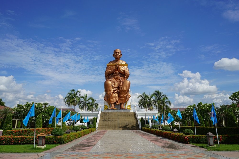 รูปปั้นทองขนาดใหญ่ของพระสงฆ์นั่งสมาธิ เที่ยวใกล้กรุงเทพ ตั้งอยู่ตรงกลางบนแท่น ล้อมรอบด้วยธง ต้นปาล์ม และสวนที่จัดแต่งอย่างสวยงามภายใต้ท้องฟ้าที่มีเมฆบางส่วน—เป็นหนึ่งในไฮไลท์ของสถานที่ท่องเที่ยวในจังหวัดสตูล.