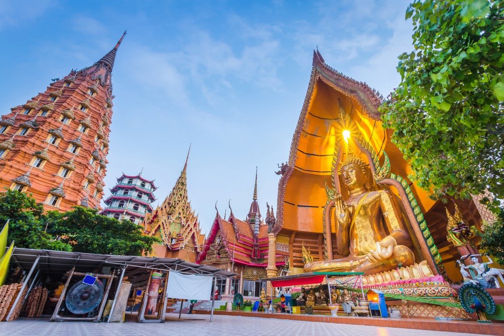 พระพุทธรูปทองคำขนาดใหญ่ประทับนั่งกลางแจ้งที่วัดถ้ำเสือใน ที่เที่ยวใกล้กรุงเทพ ประเทศไทย พร้อมด้วยอาคารวัดที่ประดับประดาอย่างวิจิตรและยอดแหลมภายใต้ท้องฟ้าสีฟ้าสดใส—เหมาะสำหรับผู้ที่มองหาทัศนียภาพที่น่าประทับใจนอกเหนือจากสถานที่ท่องเที่ยวสตูล.
