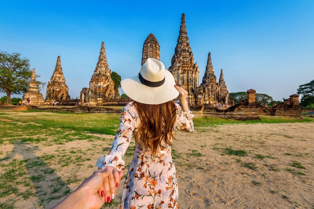 ผู้หญิงในชุดเดรสและหมวกถือมือของ ที่เที่ยวใกล้กรุงเทพธรรมชาติ ใครบางคนขณะเดินไปยังซากปรักหักพังของวัดโบราณที่มียอดแหลมสูงในวันที่มีแดดสดใส ถ่ายทอดเสน่ห์ของสถานที่ท่องเที่ยวในจังหวัดสตูล.