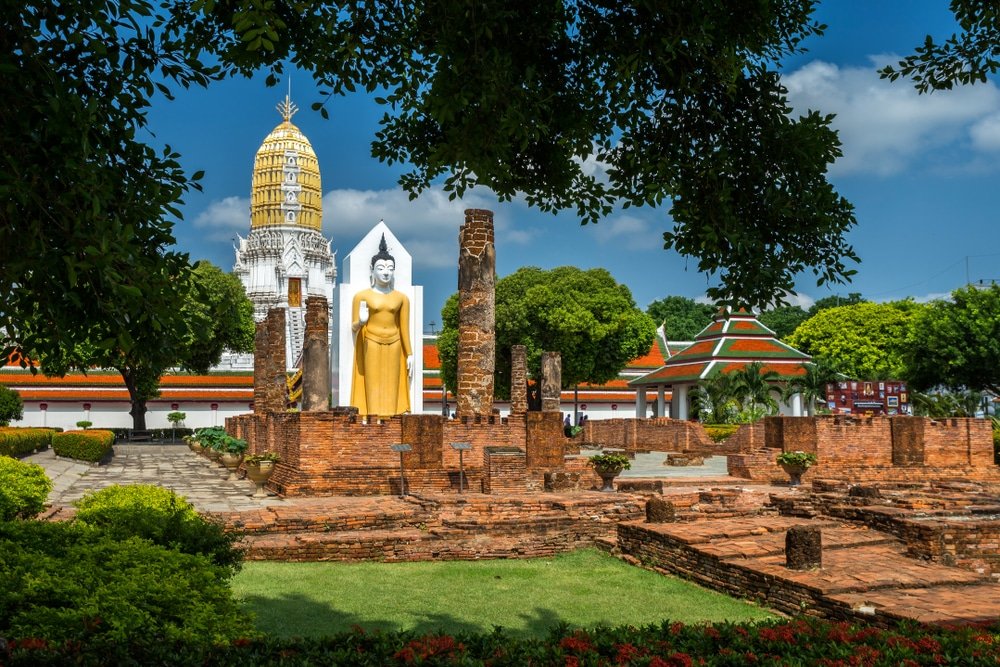 วัดโบราณในสถานที่ท่องเที่ยวของจังหวัดสตูล มีพระพุทธรูปยืนขนาด เที่ยวใกล้กรุงเทพ ใหญ่ โครงสร้างอิฐที่พังทลาย และเจดีย์สีขาวและทองตั้งอยู่ท่ามกลางต้นไม้เขียวขจีและท้องฟ้าสีคราม.