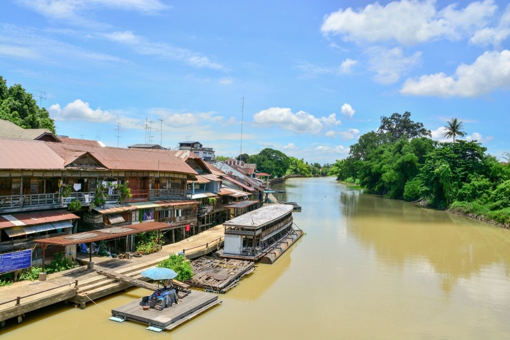 แม่น้ำที่ขนาบข้างด้วยบ้านไม้และพืชพรรณเขียวขจีในวันที่แดดจ้า พร้อม ที่เที่ยวใกล้กรุงเทพธรรมชาติ ด้วยแพลอยน้ำและเรือจอดอยู่ใกล้ริมฝั่ง—เหมาะอย่างยิ่งสำหรับผู้ที่กำลังค้นหาสถานที่ท่องเที่ยวสตูลที่เต็มไปด้วยเสน่ห์และความงดงามของธรรมชาติ.
