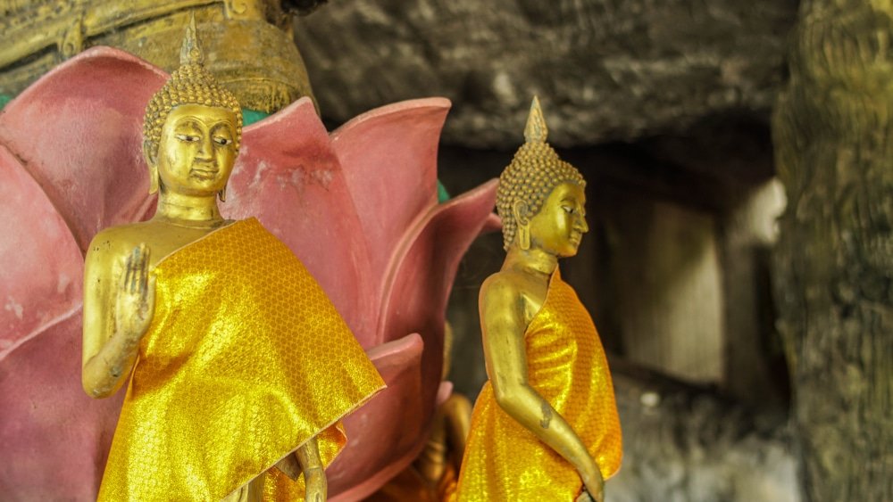 พระพุทธรูปทองคำสององค์ที่สวมจีวร ท่องเที่ยวหัวหิน สีส้มยืนอยู่ต่อหน้ารูปปั้นดอกบัวสีชมพูขนาดใหญ่ในสภาพแวดล้อมคล้ายถ้ำที่มีแสงสลัว มอบมุมมองอันเงียบสงบของสถานที่ท่องเที่ยวหัวหินสำหรับผู้ที่สำรวจจุดหมายปลายทางอันเงียบสงบแห่งนี้.