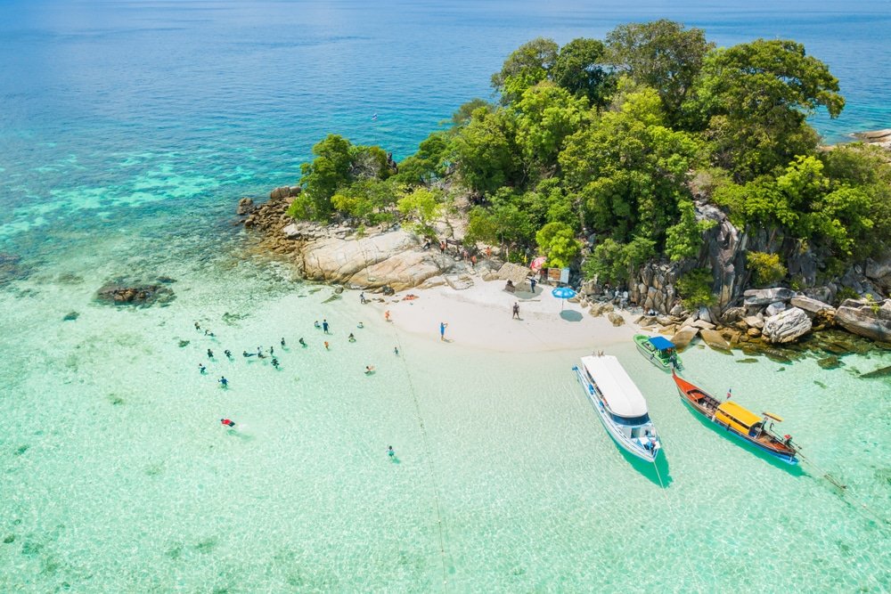 ภาพถ่ายทางอากาศของเกาะเขตร้อนขนาดเล็กใกล้หัวหิน มีน้ำทะเลสีฟ้าใส ผู้คนกำลังว่ายน้ำและดำน้ำดูปะการัง ท่องเที่ยวสตูล เรือจอดทอดสมออยู่ใกล้ชายหาดทราย—เหมาะสำหรับผู้ที่ชื่นชอบการท่องเที่ยวธรรมชาติในหัวหิน.