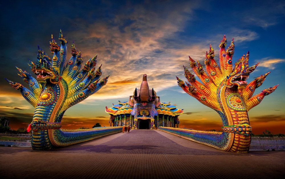 ทางเข้าวัดสีสันสดใส ประดับด้วยรูปปั้นงูขนาดใหญ่และวิจิตร พร้อม ที่เที่ยวใกล้กรุงเทพ ลวดลายแกะสลักที่ซับซ้อน ท่ามกลางพระอาทิตย์ตกดินบนท้องฟ้าที่งดงามตระการตา—สถานที่ท่องเที่ยวที่ไม่ควรพลาดในสตูล สำหรับผู้ที่กำลังค้นหาความงามของภาคใต้ของประเทศไทย.