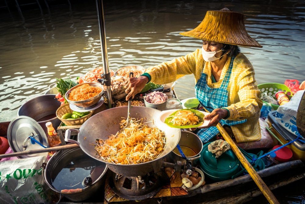 พ่อค้าในหมวกฟางและหน้ากากกำลังปรุงและเสิร์ฟอาหารจากเรือในตลาด เที่ยวใกล้กรุงเทพ น้ำ ล้อมรอบด้วยวัตถุดิบและเครื่องครัวหลากหลายชนิด—เป็นภาพที่พบได้บ่อยในแหล่งท่องเที่ยวสตูล.