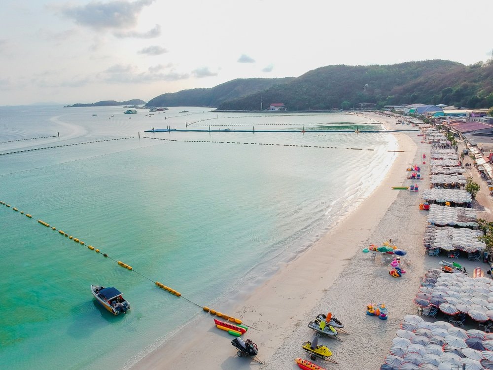 ภาพมุมสูงของชายหาดทรายขาวสะอาด น้ำทะเลสีเทอร์ควอยซ์ใส ที่เที่ยวใกล้กรุงเทพ สะอาด เรือ, เจ็ตสกี, และร่มกับเก้าอี้อาบแดดเรียงราย; ภูเขาและตึกในพื้นหลัง—สถานที่ท่องเที่ยวสตูลที่สวยงามสำหรับทริปต่อไปของคุณ.