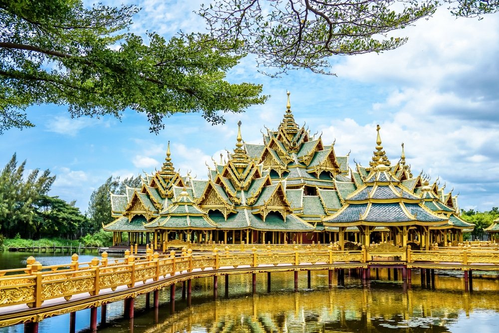 ศาลาไทยแบบดั้งเดิมที่มีหลังคา ที่เที่ยวใกล้กทม ทองคำประดับอย่างวิจิตรตั้งอยู่เหนือผืนน้ำ เชื่อมต่อด้วยทางเดินที่ประณีต ภายใต้ท้องฟ้าที่มีเมฆบางส่วน—เป็นภาพที่สงบเงียบชวนให้นึกถึง สถานที่ท่องเที่ยวในจังหวัดสตูล.