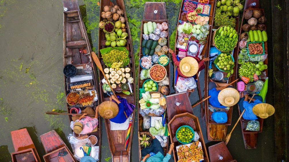 ภาพมุมสูงของเรือไม้ที่บรรทุกผลไม้ เที่ยวใกล้กรุงเทพ ผัก และสินค้าต่างๆ ในตลาดน้ำ พร้อมพ่อค้าแม่ค้าที่สวมหมวกฟาง—เป็นภาพที่เต็มไปด้วยชีวิตชีวา สะท้อนเสน่ห์ของการท่องเที่ยวสตูลและบรรยากาศอันเป็นเอกลักษณ์ของสถานที่ท่องเที่ยวในจังหวัดสตูล.