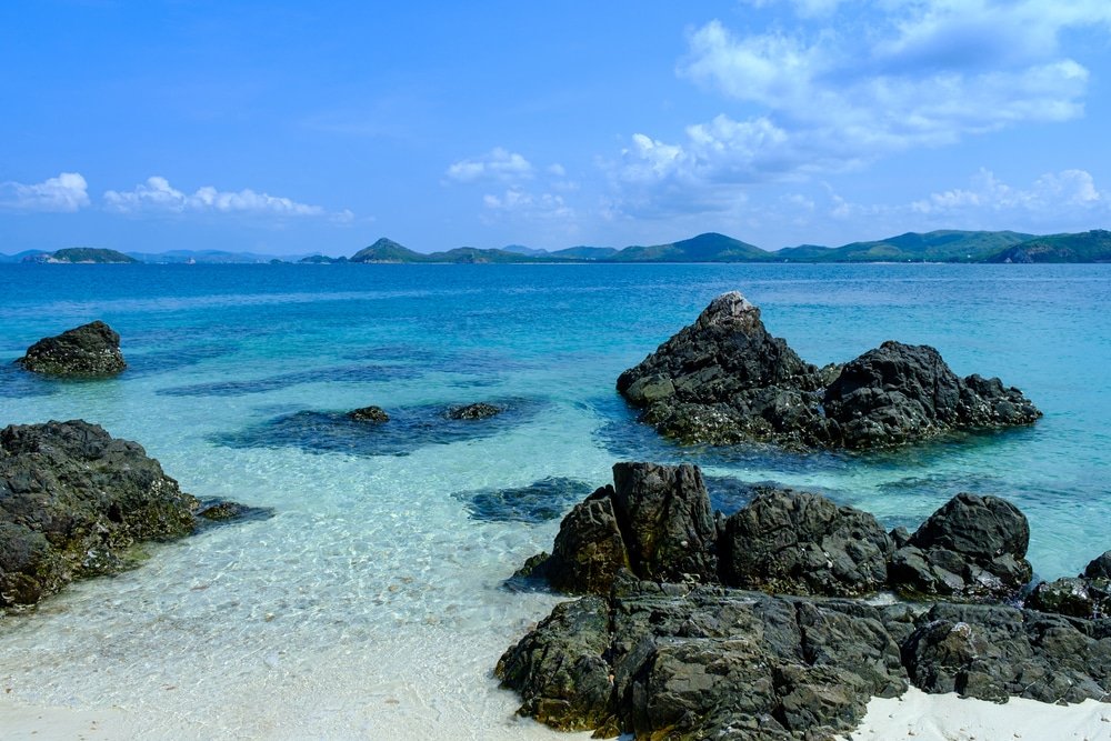 ทะเลสีฟ้าใสกับหินสีดำกระจายอยู่ใกล้ ที่เที่ยวใกล้กรุงเทพ ชายฝั่ง ชายหาดทรายขาวในเบื้องหน้า และเนินเขาสีเขียวไกลโพ้นใต้ท้องฟ้าที่มีเมฆบางส่วน—อัญมณีที่เงียบสงบในหมู่สถานที่ท่องเที่ยวสตูล.