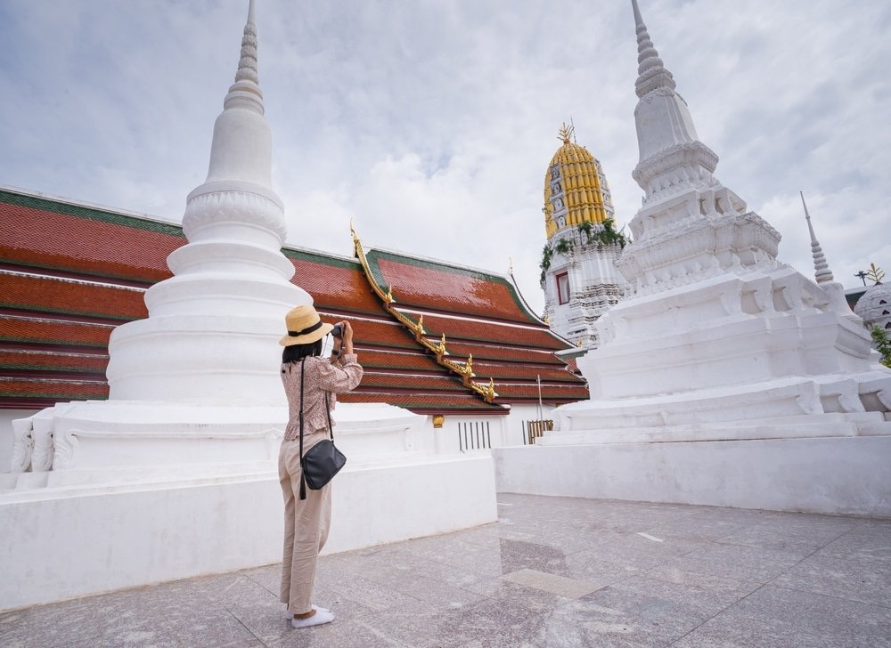 บุคคลที่สวมหมวกถ่ายภาพสถูปสีขาว เที่ยวใกล้กรุงเทพ และวัดที่มีหลังคาสีแดงและรายละเอียดสีทองภายใต้ท้องฟ้าที่มีเมฆครึ้ม จับภาพเสน่ห์ของ สถานที่ท่องเที่ยวจังหวัดสตูล.