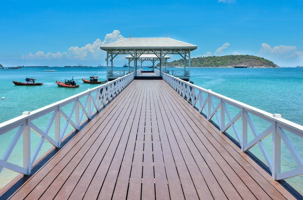 สะพานไม้ที่มีราวจับสีขาวทอดยาวเหนือผืนน้ำสีฟ้าใสไปยังศาลาที่มี ที่เที่ยวใกล้กรุงเทพ หลังคาคลุม เชิญชวนให้คุณมาค้นพบ ท่องเที่ยวสตูล พร้อมเรือและเกาะที่มองเห็นอยู่เบื้องหลังภายใต้ท้องฟ้าที่สดใส.