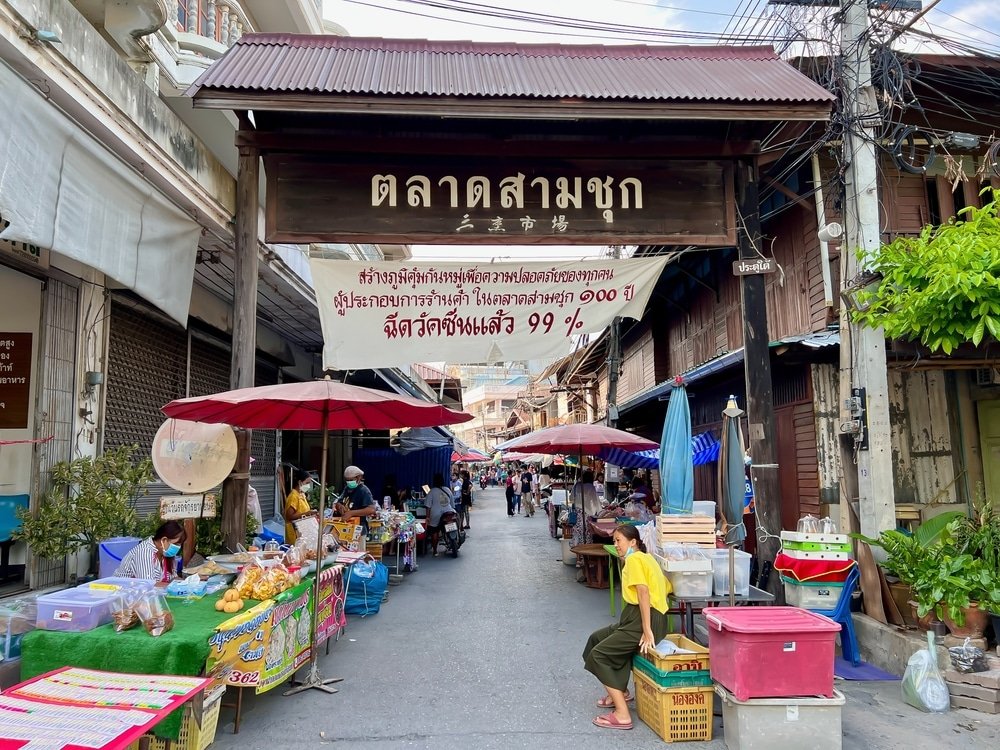 ภาพตลาดกลางแจ้งในประเทศไทยที่ ที่เที่ยวใกล้กรุงเทพธรรมชาติ มีพ่อค้าแม่ค้าขายของใต้ร่มเรียงรายตามถนนแคบๆ ผู้คนกำลังเลือกซื้อของ และมีป้ายไม้ที่มีข้อความภาษาไทยแขวนอยู่เหนือทางเข้า—ประสบการณ์การท่องเที่ยวสตูลที่แท้จริง.