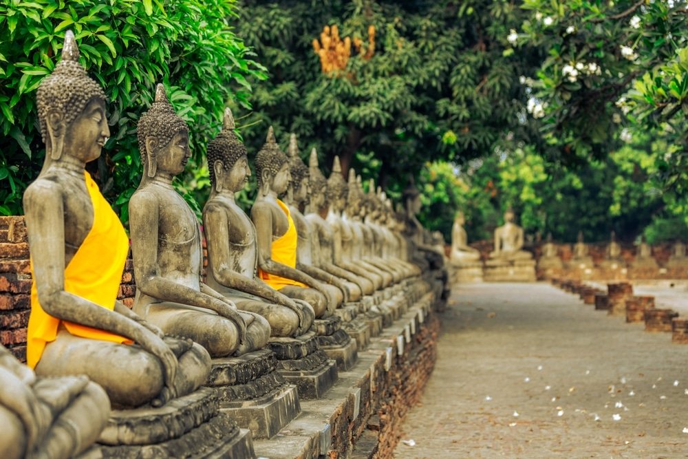 แถวของพระพุทธรูปหินที่ห่มผ้าสี ที่เที่ยวใกล้กรุงเทพธรรมชาติ เหลืองตั้งเรียงรายตามทางเดินที่ปูด้วยอิฐ ล้อมรอบด้วยพืชพรรณสีเขียว มอบภาพอันสงบเงียบที่สะท้อนถึงมรดกทางวัฒนธรรมอันล้ำค่าของสถานที่ท่องเที่ยวสตูล.