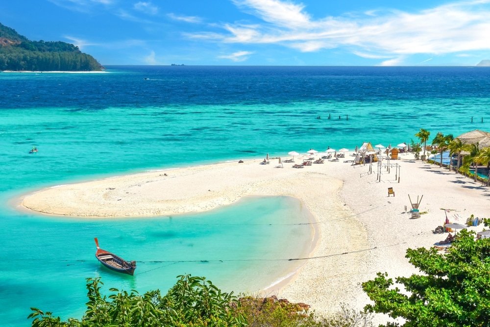 หาดทรายขาวน้ำทะเลสีฟ้าครามใส เก้าอี้นั่งเล่นและร่ม เรือไม้ในน้ำตื้น และต้นไม้เขียวขจีเบื้องหน้า เหมาะสำหรับการพักผ่อนที่เที่ยวธรรมชาติหัวหิน. ท่องเที่ยวสตูล