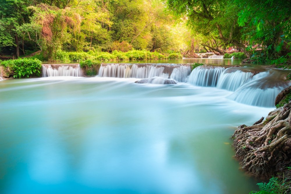 น้ำตกขนาดเล็กไหลผ่านโขดหินลงสู่แม่น้ำที่สงบเงียบ ล้อมรอบด้วยต้นไม้ ที่เที่ยวใกล้กรุงเทพ เขียวขจีและพืชพรรณที่อุดมสมบูรณ์ในแสงธรรมชาติที่สดใส—เหมาะสำหรับผู้ที่ต้องการท่องเที่ยวสตูลและสำรวจความงามของสถานที่ท่องเที่ยวในจังหวัดสตูล.
