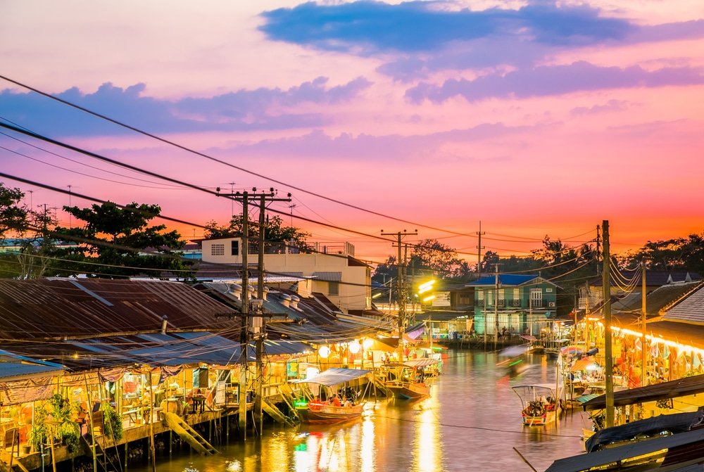 ตลาดน้ำที่เต็มไปด้วยสีสันสดใส มีเรือและแผงลอยที่ประดับไฟตามแนว เที่ยวใกล้กรุงเทพ คลองในช่วงพระอาทิตย์ตก ท่ามกลางท้องฟ้าที่สดใส มีเมฆและสายไฟฟ้าที่มองเห็นได้—เป็นภาพที่น่าหลงใหลที่มักพบได้ที่ ท่องเที่ยวสตูล สำหรับผู้ที่ชื่นชอบการสำรวจตลาดที่ไม่เหมือนใคร.