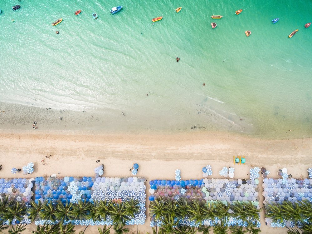 ภาพมุมสูงของชายหาดทรายขาว มีร่ม ที่เที่ยวใกล้กรุงเทพ เรียงรายเป็นแถว ต้นปาล์ม และผู้คนกำลังว่ายน้ำในน้ำทะเลสีฟ้าใสพร้อมเรือลำน้อยอยู่ใกล้ๆ—เป็นหนึ่งในสถานที่ท่องเที่ยวสตูลยอดนิยมสำหรับการพักผ่อนริมทะเลที่ไม่อาจลืมเลือน.