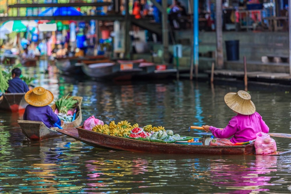 สองคนสวมหมวกพื้นเมืองพายเรือที่ เที่ยวใกล้กรุงเทพ เต็มไปด้วยผลไม้และผักไปตามคลองในตลาดน้ำลอยน้ำ บรรยากาศที่สดใสชวนให้นึกถึง สถานที่ท่องเที่ยวในจังหวัดสตูล โดยมีร้านค้าและเรือลำอื่น ๆ เป็นฉากหลัง.