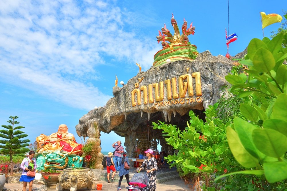 นักท่องเที่ยวเยี่ยมชมทางเข้าวัดถ้ำในประเทศไทย ซึ่งเป็นหนึ่งในไฮไลท์ของท่องเที่ยวหัวหิน โดยมีรูปปั้น ป้ายอักษรไทย และธงชาติใต้ท้องฟ้าสีคราม. ท่องเที่ยวหัวหิน 