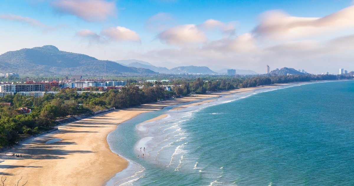 ภูมิทัศน์ชายฝั่งทะเลที่มีหาดทราย ท่องเที่ยวหัวหิน คลื่นอ่อนโยน แนวอาคารและต้นไม้ตามแนวชายฝั่ง มีเนินเขาห่างไกลอยู่ใต้ท้องฟ้าที่มีเมฆเป็นบางส่วน เหมาะอย่างยิ่งสำหรับการสำรวจพื้นที่ท่องเที่ยวหัวหิน.