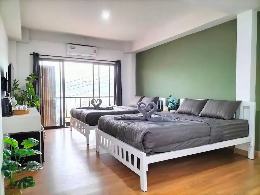 ห้องนอนสมัยใหม่ที่มีเตียงสองเตียงปูด้วยผ้าปูที่นอนสีเทา ผนังสีเขียวเป็นจุดเด่น พื้นไม้ ต้นไม้ในกระถาง และประตูบานเลื่อนกระจกที่นำไปสู่ เที่ยวหน้าฝนโรแมนติก ระเบียง ผ้าเช็ดตัวพับเป็นรูปหัวใจวางอยู่บนเตียงแต่ละเตียง.