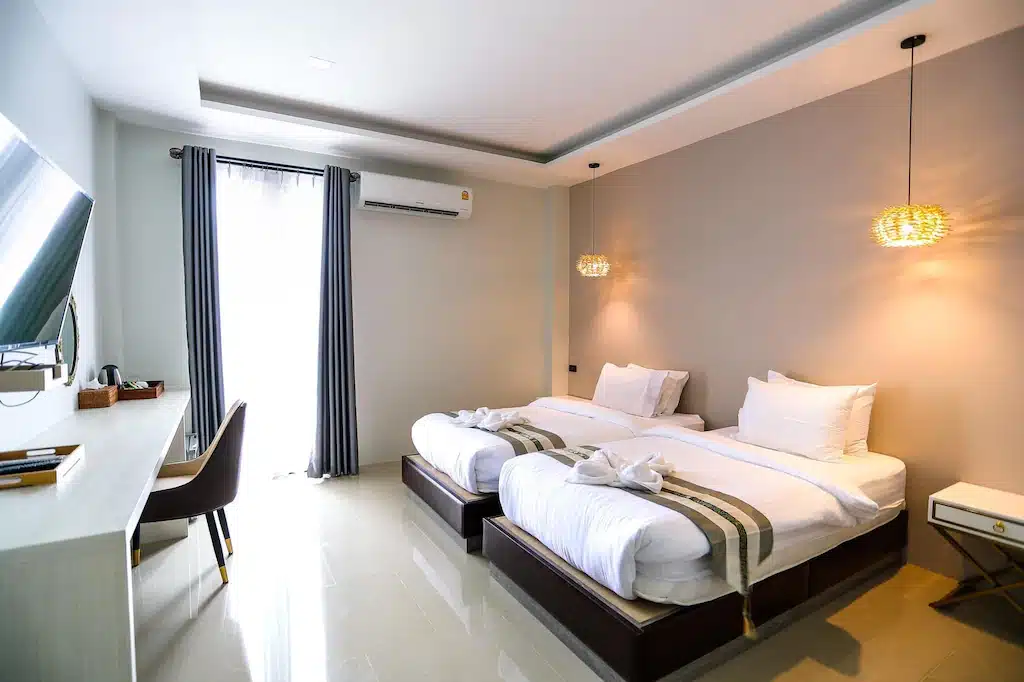 ห้องพักโรงแรมทันสมัยพร้อมเตียงเดี่ยวสองเตียง โต๊ะและเก้าอี้ ทีวีติดผนัง เครื่องปรับอากาศ และโคมไฟแขวน ผ้าเช็ดตัวพับวางไว้บนเตียงแต่ละเตียง—เหมาะสำหรับผู้ที่มองหา ที่พักภูทับเบิก เพื่อการพักผ่อนที่สะดวกสบาย.