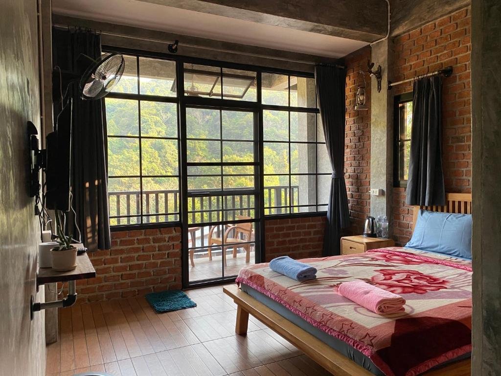ห้องนอนที่มีหน้าต่างบานใหญ่และ ที่เที่ยวหน้าฝนโรแมนติก ระเบียง พร้อมเตียงคู่ที่มีผ้าห่มสีสันสดใส ผนังอิฐ พื้นไม้ และวิวต้นไม้เขียวขจีด้านนอก.