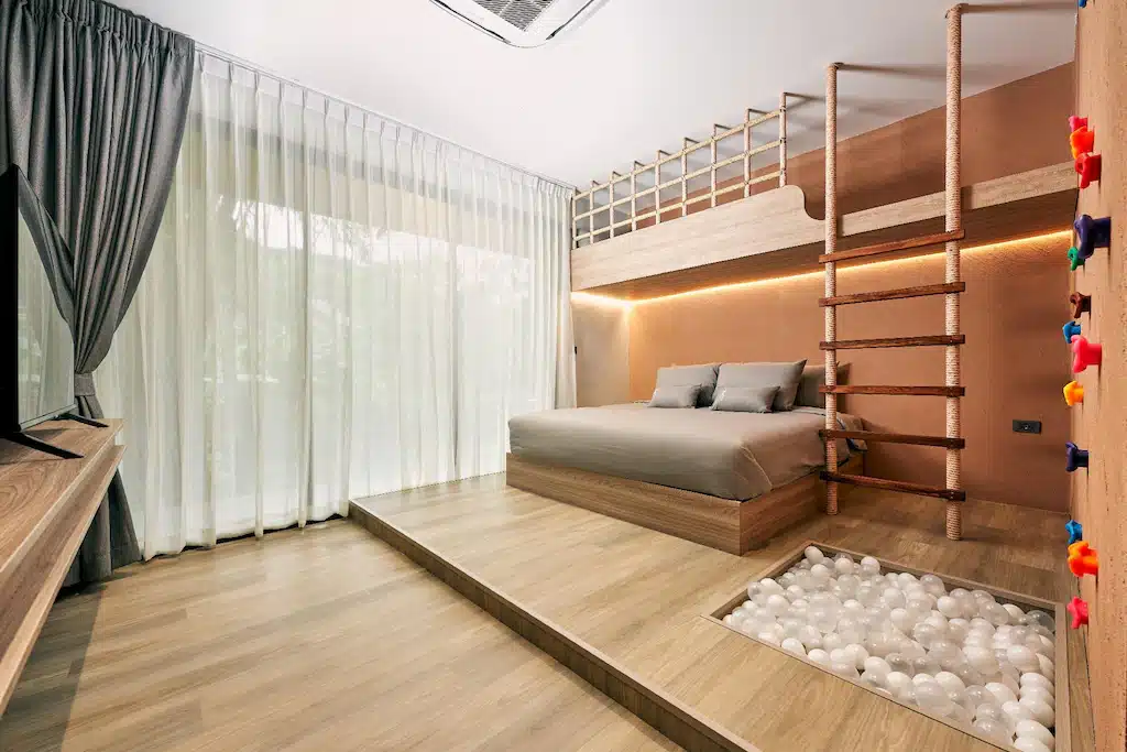 ห้องนอนสมัยใหม่พร้อมเตียงยกสูง ชั้นลอยพร้อมบันไดเชือก ที่จับปีนผนัง ทีวี หน้าต่างบานใหญ่พร้อมผ้าม่านบาง และบ่อบอลในตัว—เหมาะสำหรับครอบครัวที่มองหาประสบการณ์ รีสอร์ทภูทับเบิก ที่ไม่เหมือนใคร.
