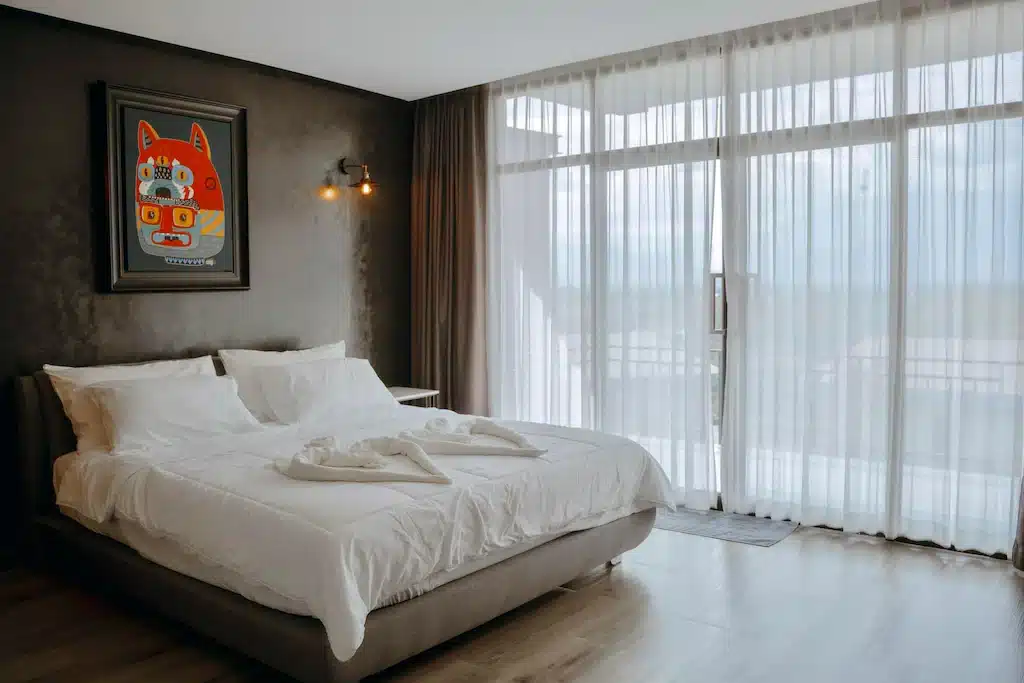ห้องนอนสมัยใหม่ที่ รีสอร์ทภูทับเบิก พร้อมเตียงขนาดใหญ่ ผ้าปูที่นอนสีขาว ผ้าเช็ดตัวพับเป็นรูปหงส์ หน้าต่างบานใหญ่จากพื้นจรดเพดานพร้อมผ้าม่านบางเบา และภาพวาดสีสันสดใสติดผนัง.