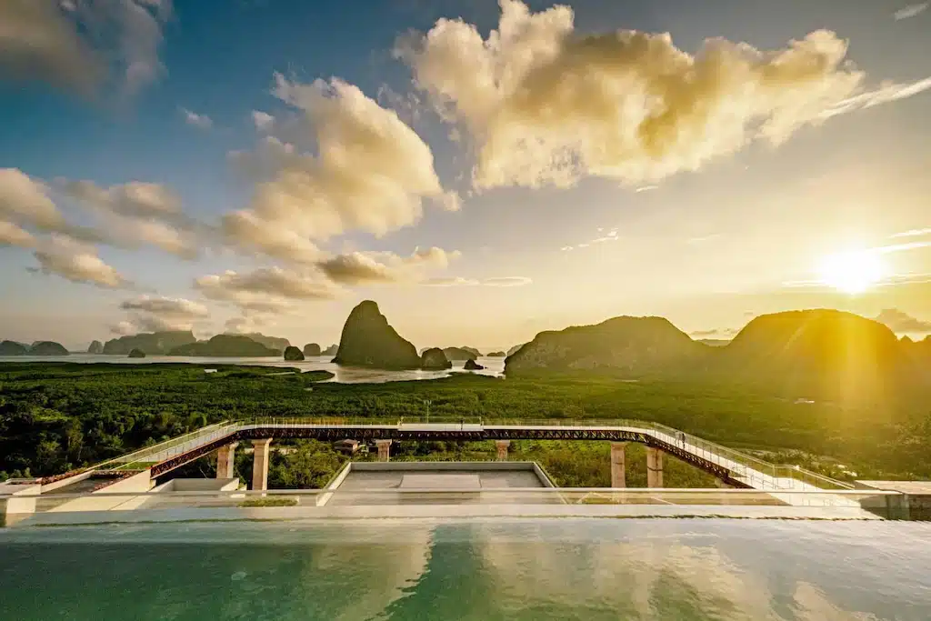 สระว่ายน้ำไร้ขอบพร้อมวิวทิวทัศน์ของภูมิทัศน์สีเขียวชอุ่ม เกาะหินปูน และ ที่เที่ยวหน้าฝนโรแมนติก พระอาทิตย์ขึ้นหรือตกดินหลังภูเขาไกลโพ้นภายใต้ท้องฟ้าที่มีเมฆบางส่วน.