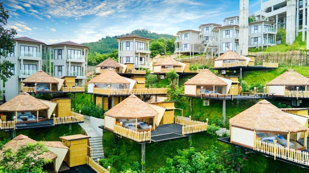 รีสอร์ทบนเนินเขาที่มีอาคารสีขาวทันสมัยและกระท่อมหลังคามุงจากหลาย ที่เที่ยวหน้าฝนโรแมนติก หลังบนเสาสูง ล้อมรอบด้วยพื้นที่สีเขียวภายใต้ท้องฟ้าที่มีเมฆบางส่วน.