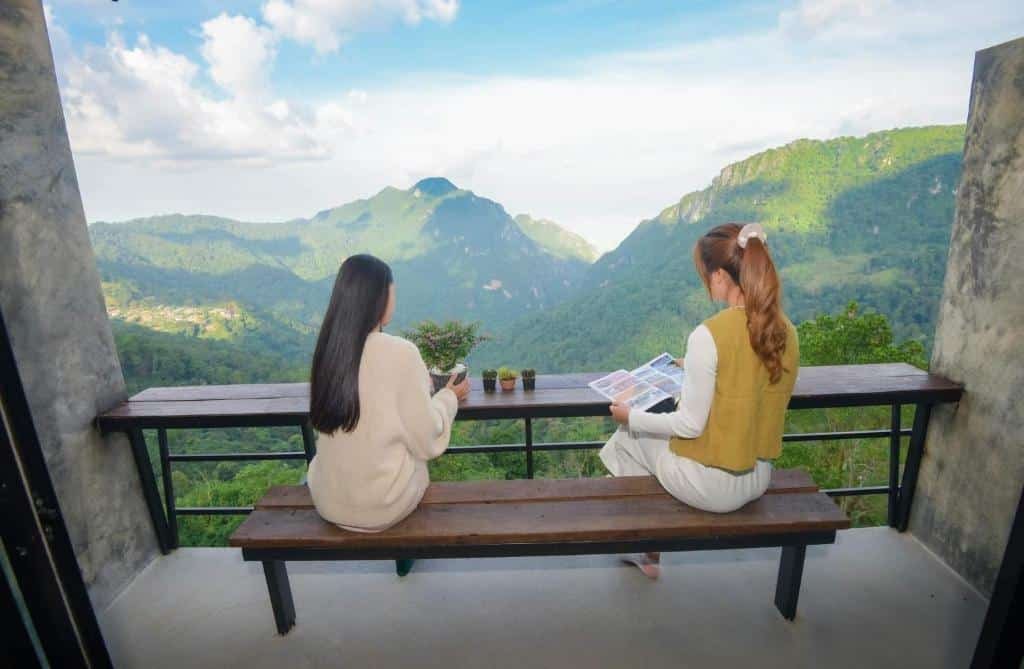 ผู้หญิงสองคนนั่งอยู่บนม้านั่งหันหน้าไปทางวิวภูเขาที่สวยงาม; คนหนึ่งมองดู ที่เที่ยวหน้าฝนโรแมนติก ทิวทัศน์ในขณะที่อีกคนหนึ่งอ่านหนังสือหรือนิตยสาร.