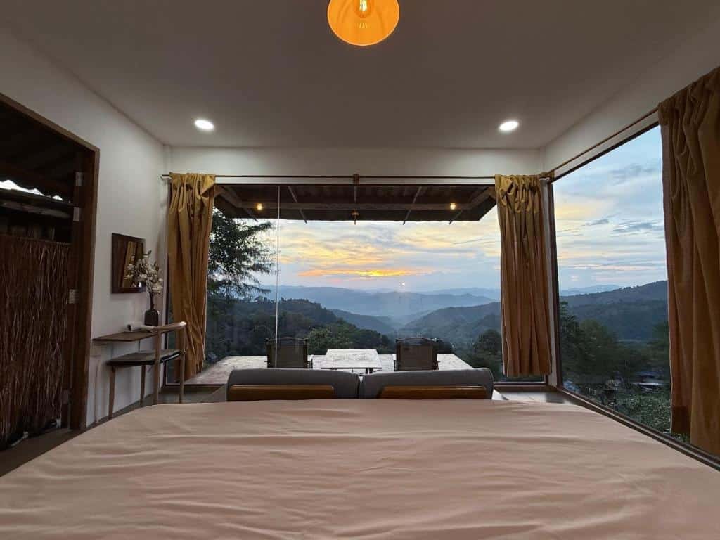 ห้องนอนพร้อมหน้าต่างบานใหญ่ ที่เที่ยวหน้าฝนโรแมนติก และผ้าม่าน ให้ทัศนียภาพของภูเขาและท้องฟ้าในยามพระอาทิตย์ตกดิน พร้อมเก้าอี้สองตัวและโต๊ะบนระเบียงภายนอก.