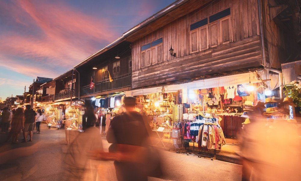 ตลาดนัดที่คึกคักในยามพลบค่ำ มีผู้คน ที่เที่ยวหน้าฝนโรแมนติก เดินผ่านไปมาอย่างเบลอๆ ผ่านอาคารไม้และร้านค้าที่สว่างไสวด้วยไฟส่องสว่าง แสดงเสื้อผ้าและสินค้าต่างๆ.