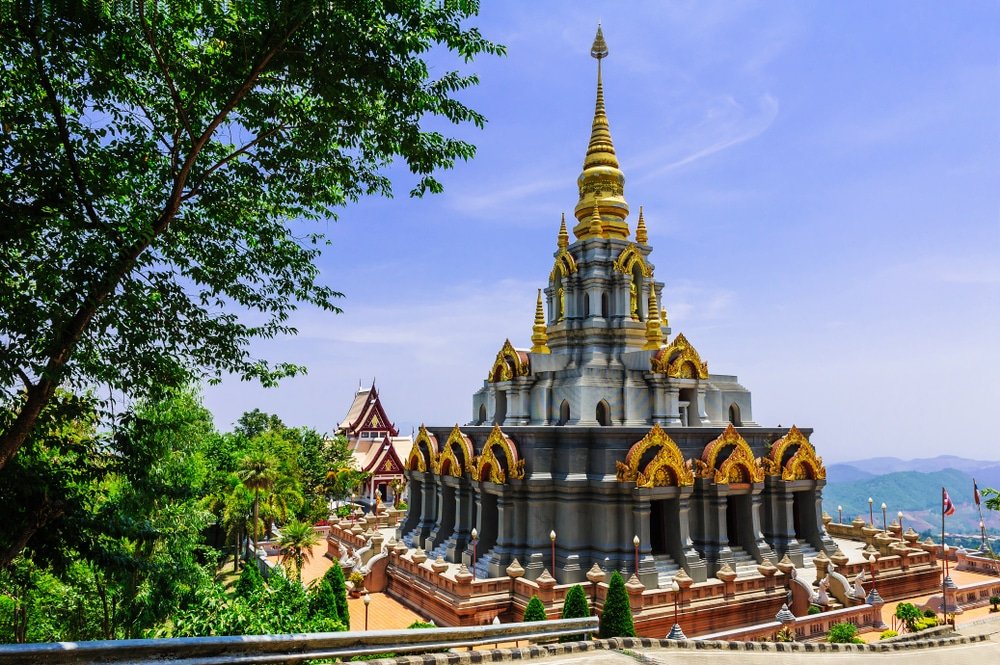 วัดพุทธที่มีรายละเอียดทองคำประดับอย่างสวยงามและยอดแหลมสูงตั้งอยู่ท่ามกลางต้นไม้และภูเขาภายใต้ท้องฟ้ เที่ยวหน้าฝนโรแมนติก สีครามแจ่มใส.