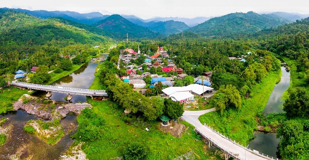 ภาพมุมสูงของหมู่บ้านเล็ก ๆ ที่ล้อมรอบด้วยเนินเขาเขียวขจี แม่น้ำ และสะพาน เที่ยวหน้าฝนโรแมนติก สองแห่งที่ทอดข้ามน้ำ.