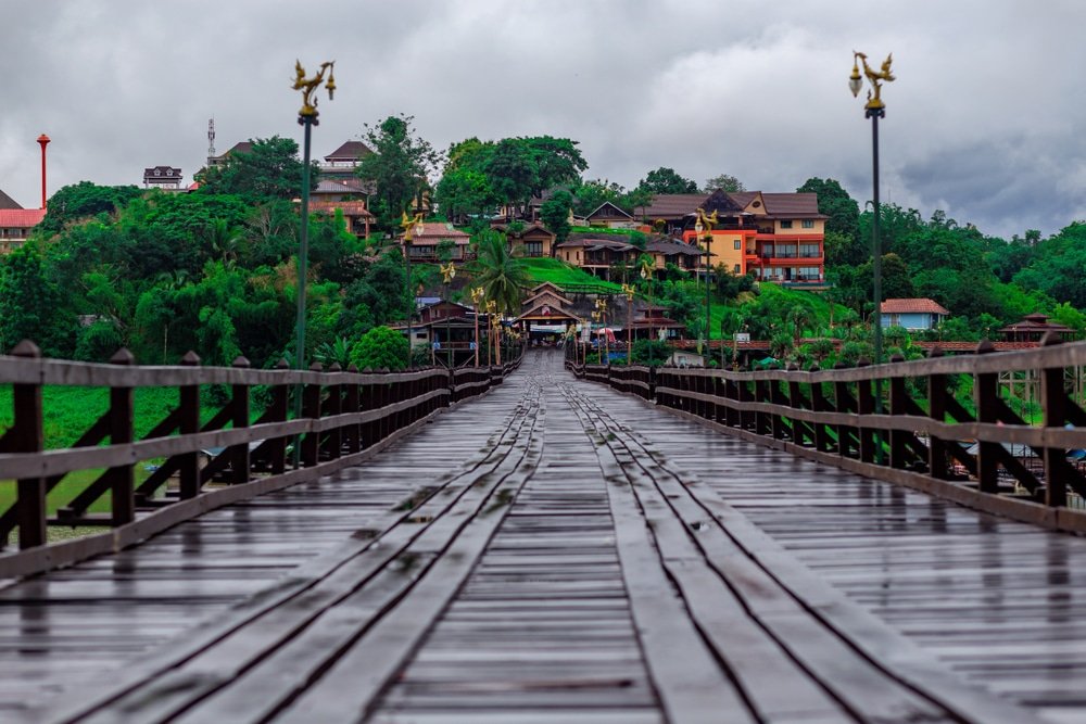 สะพานไม้เปียกชื้นทอดยาวไปยังหมู่บ้านเล็กๆ ที่มีบ้านเรือนสีสันสดใสและต้นไม้เขียวขจีอยู่ใต้ท้องฟ้าที่มีเมฆครึ้ม. เที่ยวหน้าฝนโรแมนติก 