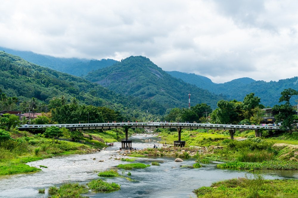 รถไฟข้ามสะพานเหนือแม่น้ำที่มีภูเขาและเนินเขาสีเขียวเป็นฉากหลัง ภายใต้ เที่ยวหน้าฝนโรแมนติก ท้องฟ้าที่มีเมฆครึ้ม.