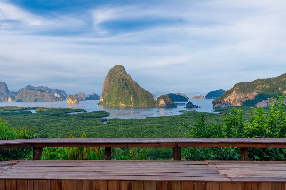 ราวไม้ทอดสายตาไปยังเกาะเขียวขจีและหน้าผาหินปูนใน ที่เที่ยวหน้าฝนโรแมนติก อ่าวที่เงียบสงบ ท่ามกลางท้องฟ้าสีครามและเมฆที่กระจายตัวอยู่เป็นระยะ.
