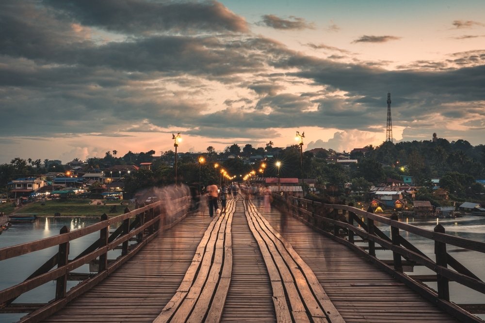 สะพานไม้กว้างที่มีผู้คนเดินข้ามในยาม เที่ยวหน้าฝนโรแมนติก พลบค่ำ ส่องสว่างด้วยแสงไฟถนน โดยมีหมู่บ้านและท้องฟ้าที่มีเมฆเป็นฉากหลัง.