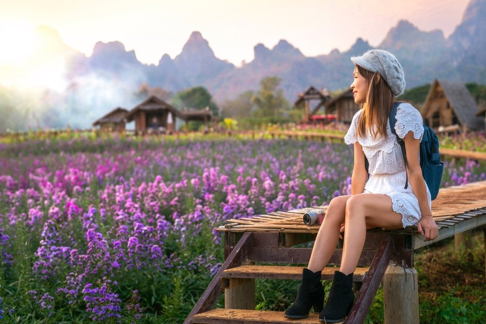 ผู้หญิงในชุดสีขาว นั่งอยู่บนบันไดไม้ มองออกไปยังทุ่งดอกไม้สีม่วง  ที่เที่ยวหน้าฝนโรแมนติก ท่ามกลางภูเขาและบ้านเรือนแบบดั้งเดิมในยามพระอาทิตย์ตกดิน.