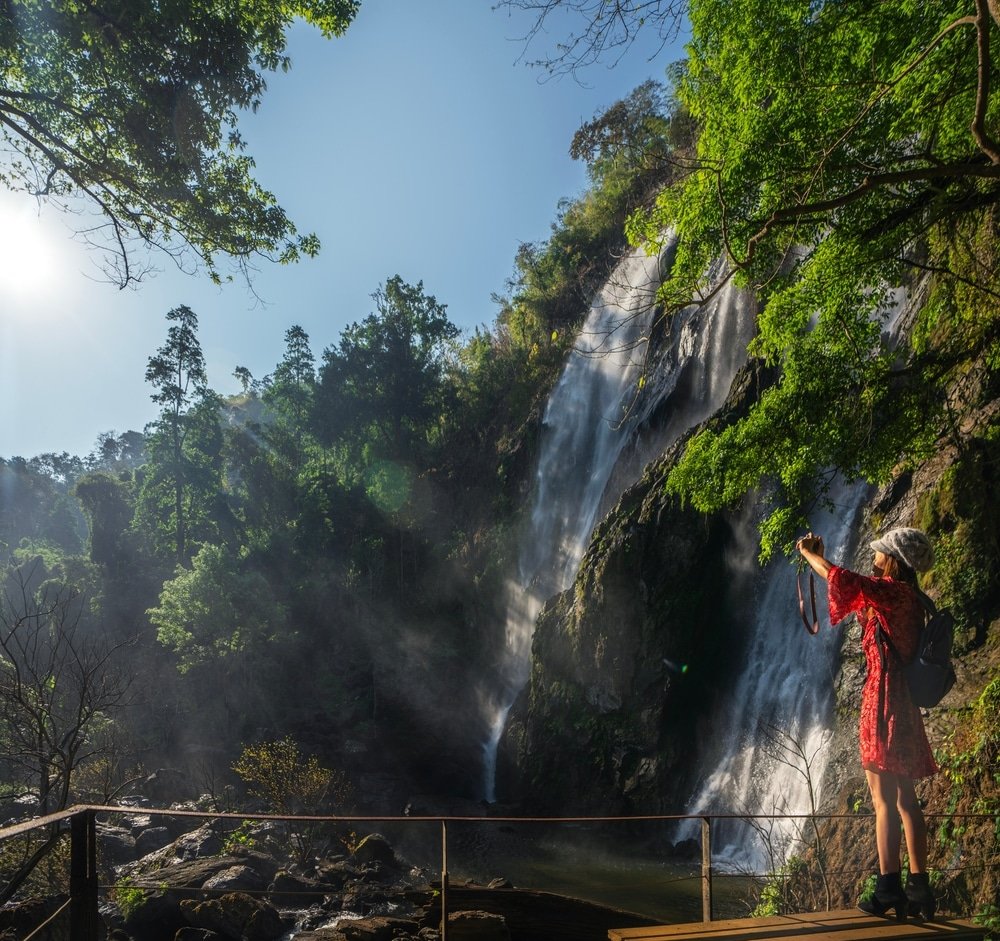 ผู้หญิงคนหนึ่งสวมชุดสีแดงยืนอยู่บน เที่ยวหน้าฝนโรแมนติก แท่นไม้ ถ่ายรูปน้ำตกที่ล้อมรอบด้วยต้นไม้สีเขียว ท่ามกลางท้องฟ้าสีครามใส.