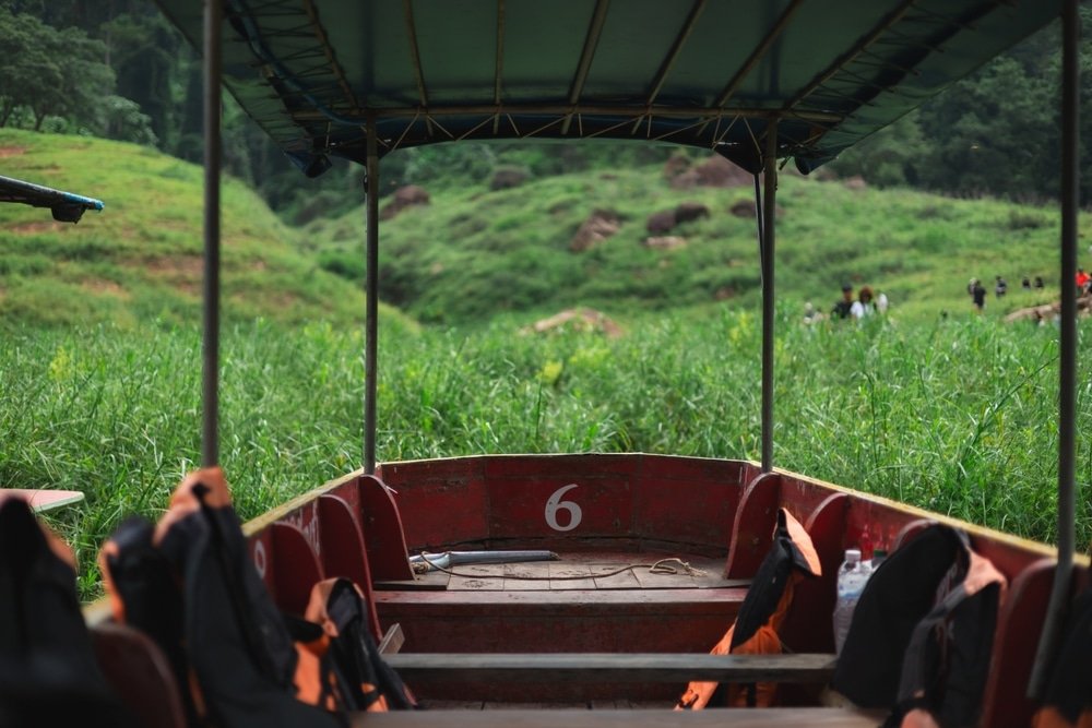 เรือสีแดงหมายเลข 6 จอดอยู่ในทุ่งหญ้าสีเขียวสูง มีที่นั่งว่างเปล่าและเสื้อชูชีพสีส้ม ล้อมรอบด้วยภูมิทัศน์ที่เต็มไปด้วย ที่เที่ยวหน้าฝนโรแมนติก หญ้า.